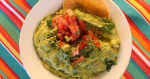 A bowl of fresh Guacamole with a chip in it on a brightly colored, striped table.