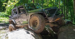 A dirty Jeep driving over some very large logs in a forest.