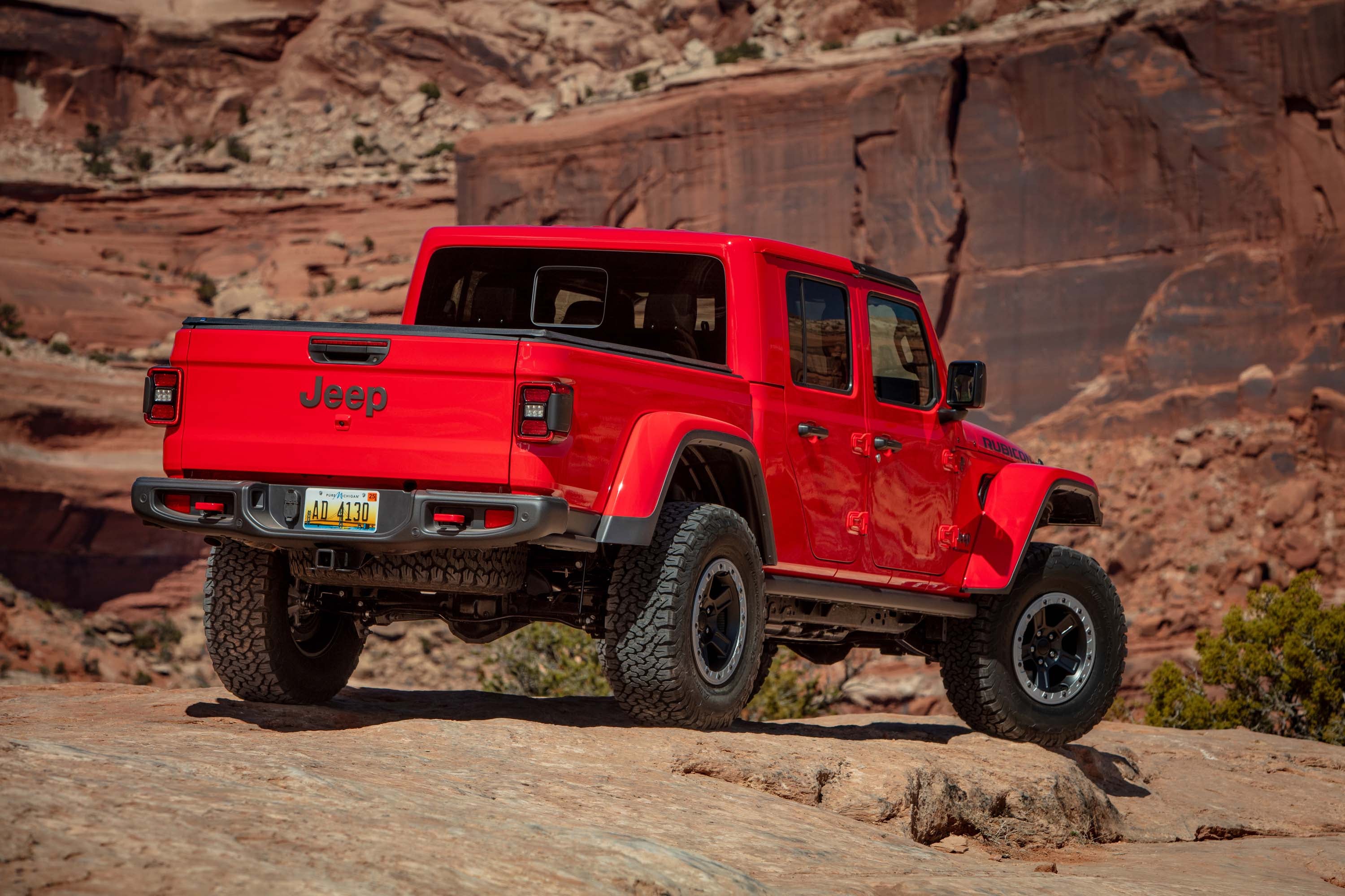 Rear three-quarter view of a red Jeep Gladiator pickup truck parked on rocky terrain with a cliff face in the background.