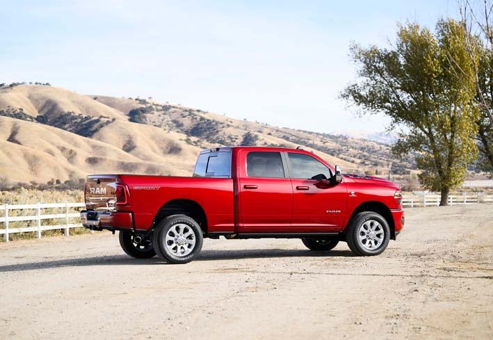 A side view of a bright red Ram 2500 Sport pickup truck is parked on a dirt road in front of rolling brown hills. There is a white fence and a single tree in the foreground. The sky is blue with no clouds.