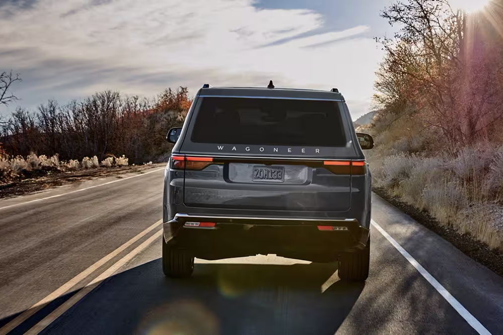 Alt text: "Rear view of a dark-colored Wagoneer SUV driving on a scenic road with double yellow lines. The vehicle features sleek LED taillights, bold badging, and a wide stance. The background includes autumn foliage, a clear sky, and the sun casting a warm glow over the landscape."