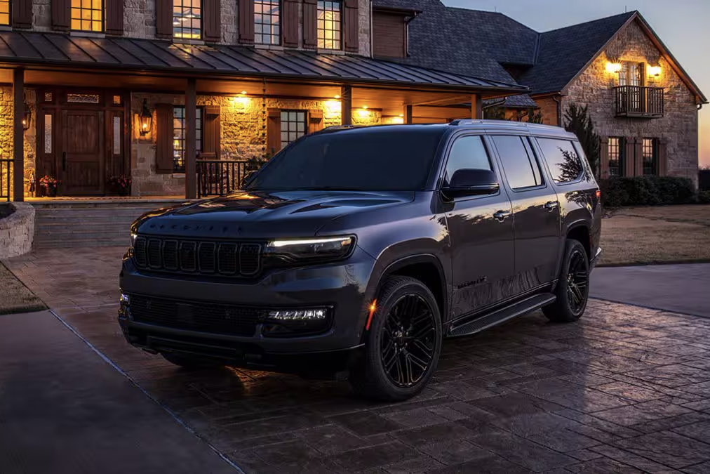 Alt text: "A black Jeep SUV with a bold front grille and sleek headlights parked on a stone driveway in front of a luxurious, well-lit home with a rustic brick and stone facade at dusk."