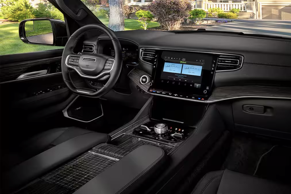 Alt text: "Luxury SUV interior featuring a black leather-wrapped steering wheel with Jeep branding, a large touchscreen infotainment system, a rotary gear selector, and premium wood and leather trim. The vehicle is parked in front of a well-manicured suburban home with a green lawn and a stone pathway."