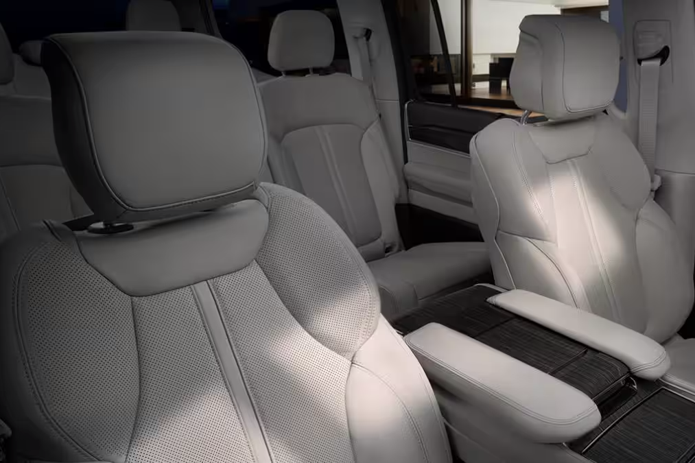 Alt text: "Luxury SUV interior showcasing plush, light-colored leather seats with perforated detailing, adjustable headrests, and a spacious cabin. The center console features wood trim and padded armrests, emphasizing comfort and premium craftsmanship."