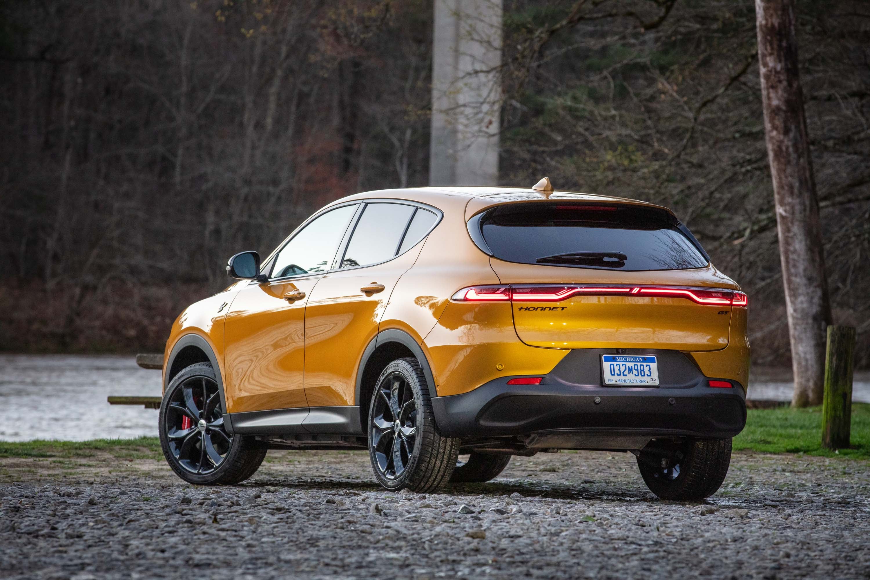 A gold-colored 2023 Dodge Hornet GT is parked on a gravel ground with its rear facing the viewer. In the background, there is a river and a forest with bare trees, and a concrete bridge column is visible on the left side.