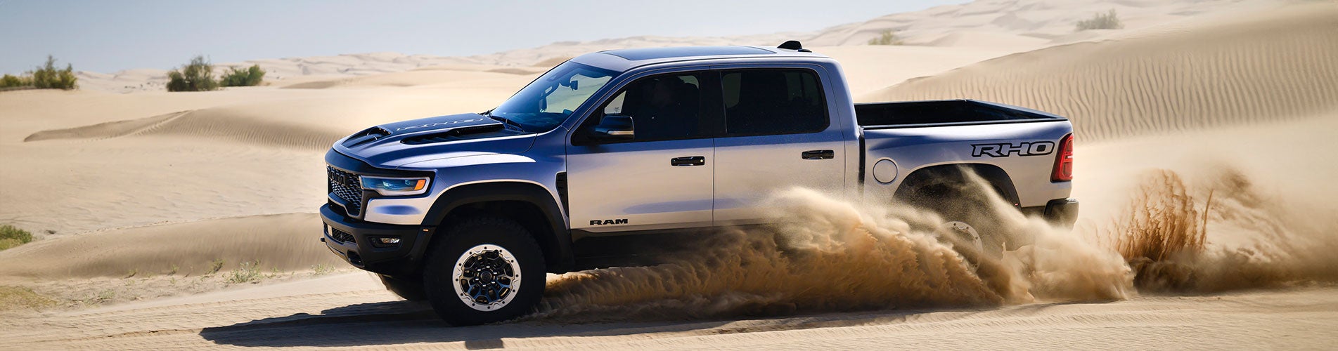 Alt text: "A silver and black RAM RHO pickup truck kicking up sand as it drives through a desert landscape. The vehicle features an aggressive front grille, black hood vents, off-road tires, and bold 'RHO' badging on the rear side. The background consists of rolling sand dunes under a clear sky."